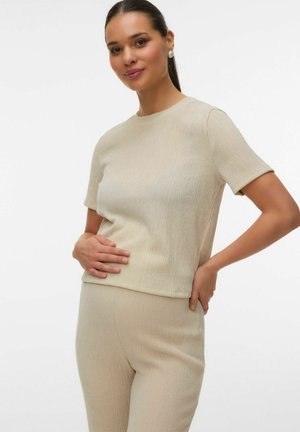 Femme aux cheveux foncés portant un haut beige à manches courtes et un pantalon assorti, touchant son ventre et souriant devant un fond blanc uni.