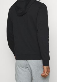 Sudadera negra con cordón y bolsillo canguro, hecha de material suave, combinada con pantalones de chándal gris claro.