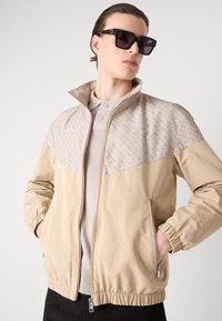 Casaco bege com fecho zip, com punhos e bainha elásticos, apresentando um painel superior texturizado em bege mais claro. O modelo veste um suéter claro por baixo do casaco.
