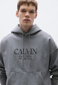Szürke kapucnis pulóver kenguruzsebbel, húzózsinóros kapucnival és fekete hímzett "CALVIN NEW YORK 1968 CK" logóval. Puha anyagtextúra.
