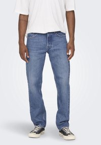 Los jeans de denim azul tienen un corte relajado, un diseño de cinco bolsillos y costuras visibles. Se llevan con una camiseta blanca y zapatillas negras.