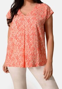 Top con stampa floreale in colori corallo e crema, caratterizzato da scollatura a V, maniche corte e pieghe per una maggiore texture. Abbinato a pantaloni chiari.