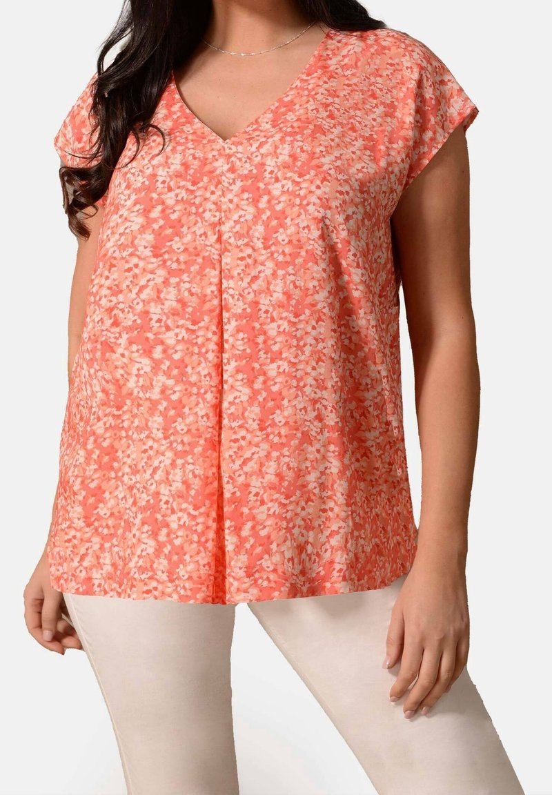 Top con stampa floreale in colori corallo e crema, caratterizzato da scollatura a V, maniche corte e pieghe per una maggiore texture. Abbinato a pantaloni chiari.