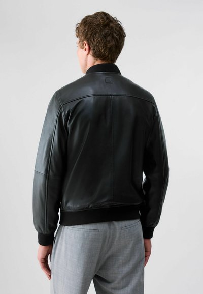 Homme portant une veste en cuir noire et un pantalon gris clair, vu de dos sur un fond clair uni.