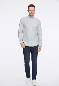 Camicia grigia chiara con bottoni e taschino sul petto, abbinata a jeans blu scuro e sneaker bianche. Texture liscia, vestibilità su misura.