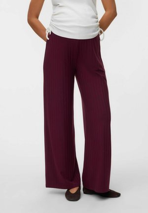 Personne debout les mains dans les poches, avec un haut blanc froncé, un pantalon bordeaux à jambes larges et des chaussures noires sur un fond uni.