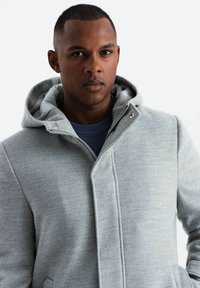 Chaqueta gris con capucha, de textura suave, con cierre de cremallera y bolsillos laterales. Cuenta con un cuello en capas y un forro interior suave.