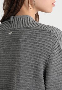Pull en maille gris à côtes avec un col montant et une petite étiquette en métal argenté à l'arrière. Le tissu semble doux et texturé.