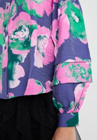 Fargerik skjorte med blomsterdesign i rosa, grønt og lilla nyanser. Teksturert stoff med knappeåpning og puffete ermer.