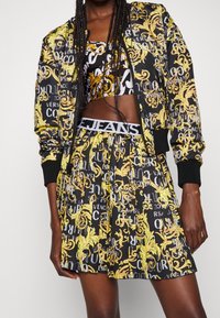 Svart bomberjacka med gul och vit grafisk text och blommönster, parat med en matchande plisserad kjol och crop top.