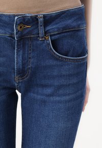 Temno modri jeans iz denima imajo kontrastno šivanje, pet žepov in gumbasto zapiranje. Gladka tekstura z rahlo obrabo.