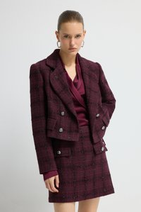 Bordeaux geruite pak met een structuurstof die doet denken aan wol, met zilverkleurige knoopaccenten en een blouse in wikkelstijl eronder.