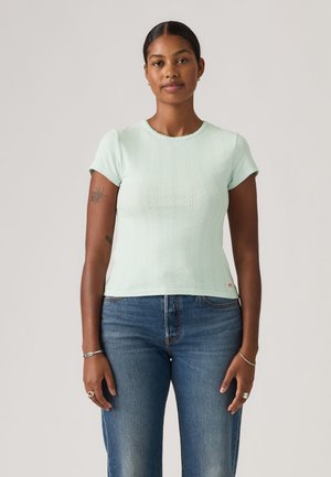 Femme aux cheveux tressés portant un haut vert clair à manches courtes et un jean bleu, debout les mains détendues le long du corps.