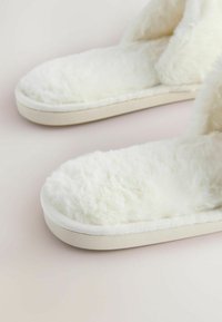 Fluffy white slippers with a soft, textured upper, and a cream-colored sole. The design features an open-toe and slip-on style.
