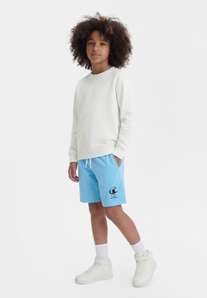 Barn med krøllete hår iført hvit genser, lyseblå shorts med logo, hvite sokker og hvite høye joggesko, står med en hånd i lommen.