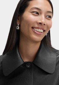 Smilende kvinde med langt mørkt hår iført sølvrunde øreringe og en sort læderjakke med en stor krave og knap.