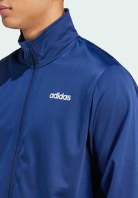 Marinblå zip-up jacka med hög krage i slätt tyg, med en vit adidas-logga på bröstet och minimalistisk design.