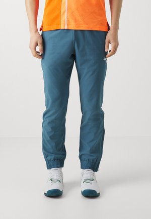 Pantaloni jogger in teal con polsini elasticizzati, materiale leggero e una sottile etichetta sul lato sinistro inferiore. Indossati con scarpe da ginnastica bianche.