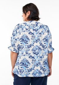 Blumenbluse aus leichtem Stoff, mit blauen und marineblauen Mustern, kurzen rüschierten Ärmeln und einem abgerundeten Saum. Die Rückansicht zeigt dekorative Details.