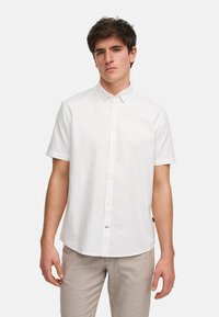 Camisa blanca de botones de manga corta hecha de tejido ligero. Presenta un cuello clásico y un corte recto, combinada con pantalones gris claro.