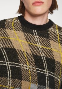 Hnědý a černý kostkovaný svetr s žlutými a bílými akcenty, s vroubkovaným černým výstřihem a strukturovanou pleteninou.