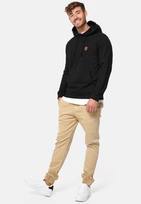 Zwarte hoodie met kangoeroezak, geribbelde boorden en een leren logopatch op de borst. Gecombineerd met beige broek en witte sneakers.