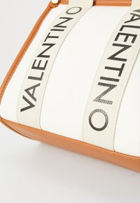 Saco de tela com corpo branco, fundo em couro castanho e alças creme, apresentando 'VALENTINO' em letras negritas pretas. Textura suave, formato retangular.