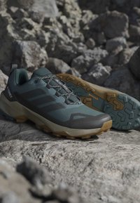 Buty sportowe z siateczkową cholewką w ciemnym odcieniu teal i szarości, gumową podeszwą z tanowym uchwytem, z czarnymi paskami i wzmocnionym noskiem.