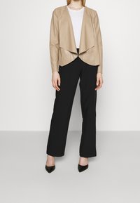 Beige koženka s otevřeným předním dílem a vrstveným designem, spárovaná s černými širokými kalhotami a špičatými černými botami.