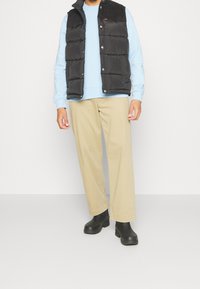 Chaleco negro acolchado con hombros de pana, sudadera azul claro, pantalones anchos beige y botines negros. Texturas suaves y ajuste casual.