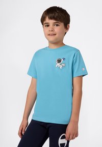 Niño vistiendo una camiseta ligera azul con un gráfico de astronauta y pantalones azul marino con un logo blanco, de pie contra un fondo plano.