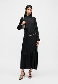 Robe noire à manches longues avec un motif texturé, un ourlet à volants et une ceinture chaîne. Le mannequin tient un petit sac à main métallique et porte des talons à brides.