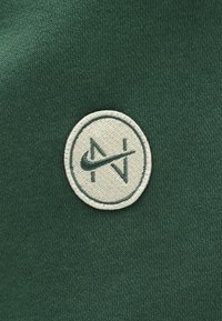 Tissu vert à texture douce présentant un logo rond brodé en écru, affichant les lettres "N" et "V" entrelacées.