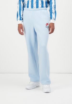 Personne portant un pantalon de survêtement Nike bleu clair avec un logo rouge et des baskets blanches, debout devant un fond blanc uni.