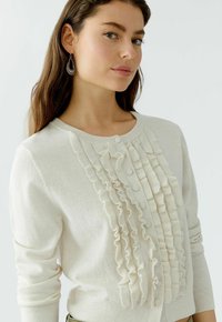 Cremfarbener Cardigan aus weichem Stoff mit einem Rüschendesign an der Vorderseite, Knöpfen, rundem Ausschnitt und langen Ärmeln.