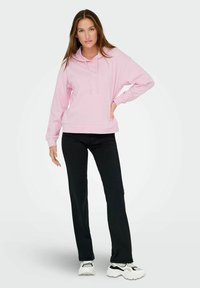 Hanorac roz deschis cu glugă și șnur, croială rectangulară și mansete din material ribbit, purtat cu pantaloni negri și adidași albi.