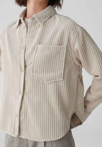 Beige, ribbed button-up shirt with a chest pocket, pointed collar, and rounded hem, made from textured fabric.