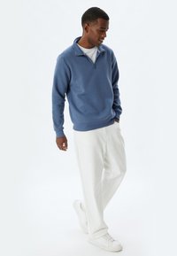 Blauer Fleece-Pullover mit einem halben Reißverschluss am Kragen, gerippten Bündchen und Saum, kombiniert mit weißen weiten Hosen und weißen Sneakers.
