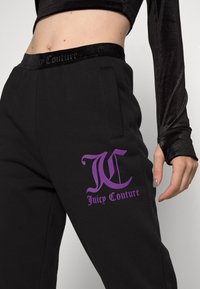 Černé tepláky vyrobené z měkké látky, s texturovaným černým pasem a fialovým logem "Juicy Couture" na levé noze.