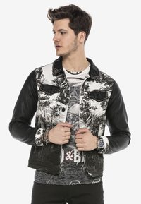 Schwarze Denimjacke mit bedrucktem weißen Stoff und Lederärmel; verfügt über gestanzte Akzente und Brusttaschen; getragen über einem gemusterten Hemd.