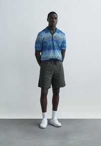 Polo de manga corta con patrón de ondas en azul y turquesa, combinado con pantalones cortos verde oliva y zapatillas blancas, sobre un fondo claro.