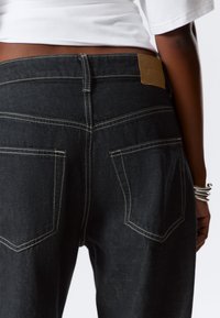 Black denim jeans with a fitted waist, contrasting white stitching, and two back pockets. A brown leather patch is attached at the back waistband.