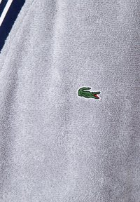 Tessuto di spugna grigio con una texture morbida, caratterizzato da un piccolo logo di coccodrillo ricamato in verde con dettagli rossi. Accenti a righe orizzontali visibili.