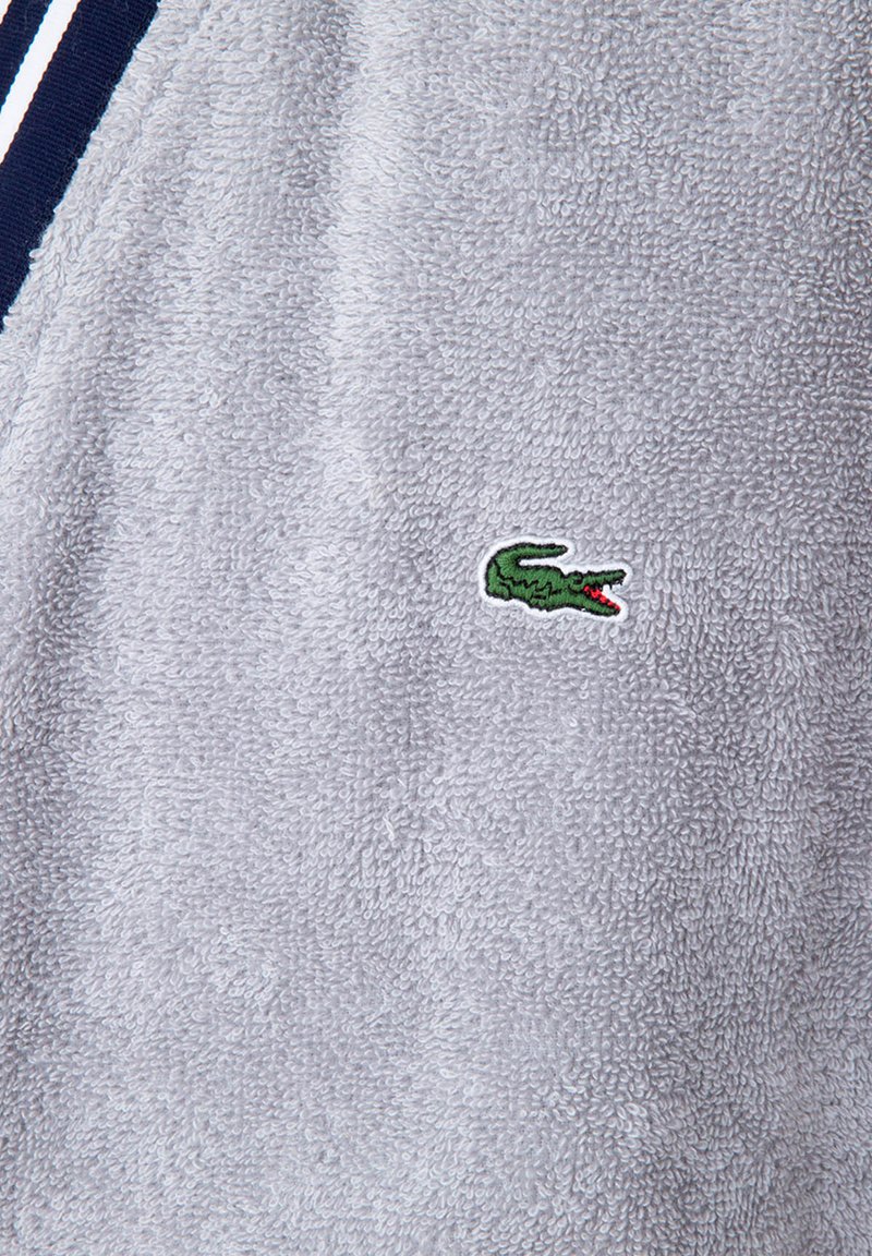 Grauer Frottee-Stoff mit weicher Textur, versehen mit einem kleinen grünen gestickten Krokodil-Logo mit roten Akzenten. Horizontale Streifen-Details sind sichtbar.