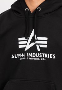 Felpa nera con cappuccio caratterizzata da un logo bianco con una lettera "A" stilizzata e tre strisce orizzontali, realizzata in tessuto morbido e dotata di chiusura con cordino.