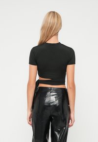 Top negru ajustat cu mâneci scurte și detaliu cu fundiță la spate, asortat cu pantaloni negri cu talie înaltă, lucioși, având o textură elegantă.