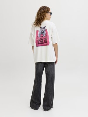 Persona con gafas y pelo rizado que lleva una camiseta blanca de gran tamaño con un gato gris y texto repetido "Thank You" sobre un fondo rosa en la parte trasera.