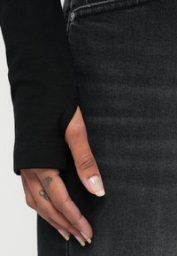 Czarna koszula z długim rękawem z otworem na kciuk i dopasowanym krojem, zestawiona z ciemnoszarymi dżinsami. Widoczna ręka z tatuażem i starannie zadbanymi paznokciami.