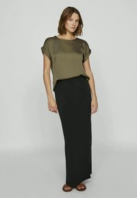 Blusa de seda verde-oliva com mangas enroçadas combinada com uma saia maxi preta ajustada. O conjunto é complementado por sandálias planas castanhas.