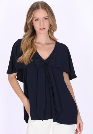 Młoda kobieta o długich blond włosach, ubrana w granatową bluzkę z kokardą i krótkimi falbankowymi rękawami, zestawioną z białymi spodniami.